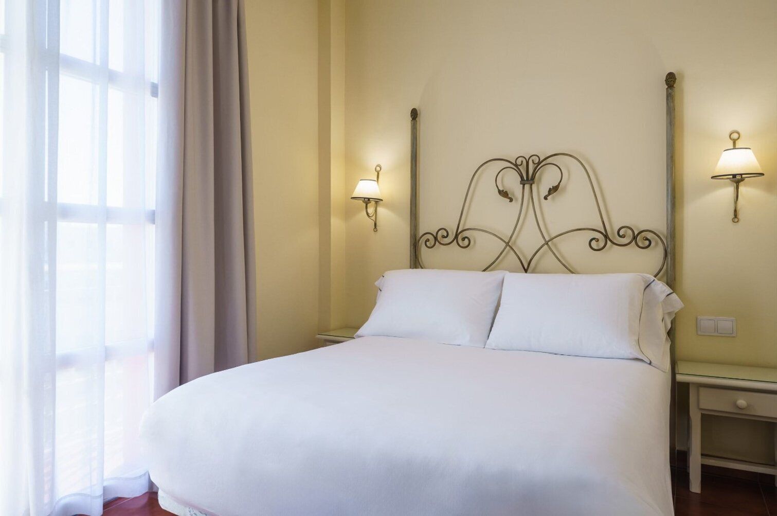 A neatly made bed in a hotel room with a simple headboard and two wall-mounted lamps.