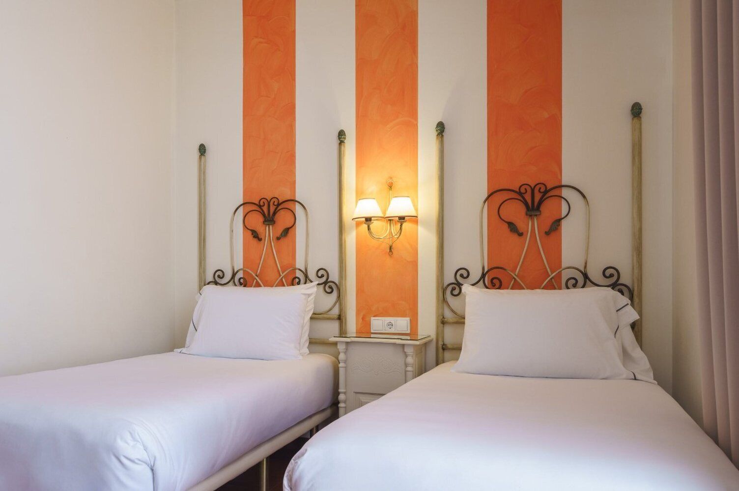 Habitación de hotel con dos camas individuales, cada una con colchón y almohada blancos. Hay una mesita de noche pequeña con una lámpara entre las camas. Las paredes presentan un diseño a rayas con paneles naranja y blanco.