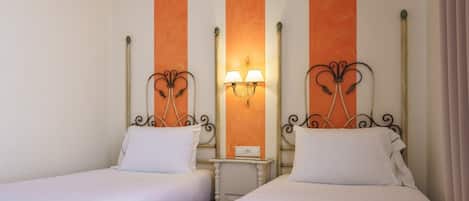 Habitación de hotel con dos camas individuales, cada una con colchón y almohada blancos. Hay una mesita de noche pequeña con una lámpara entre las camas. Las paredes presentan un diseño a rayas con paneles naranja y blanco.