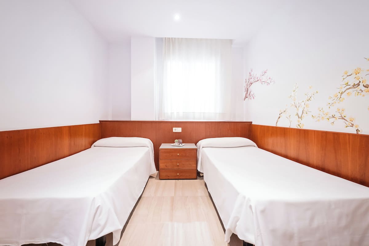 Une chambre d’hôtel avec deux lits simples, une tête de lit en bois, une petite table de chevet avec une lampe et une fenêtre avec des rideaux légers.