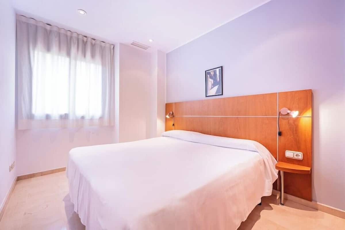 Une chambre d’hôtel avec un lit, une table de chevet, une lampe fixée au mur et une fenêtre avec des rideaux.