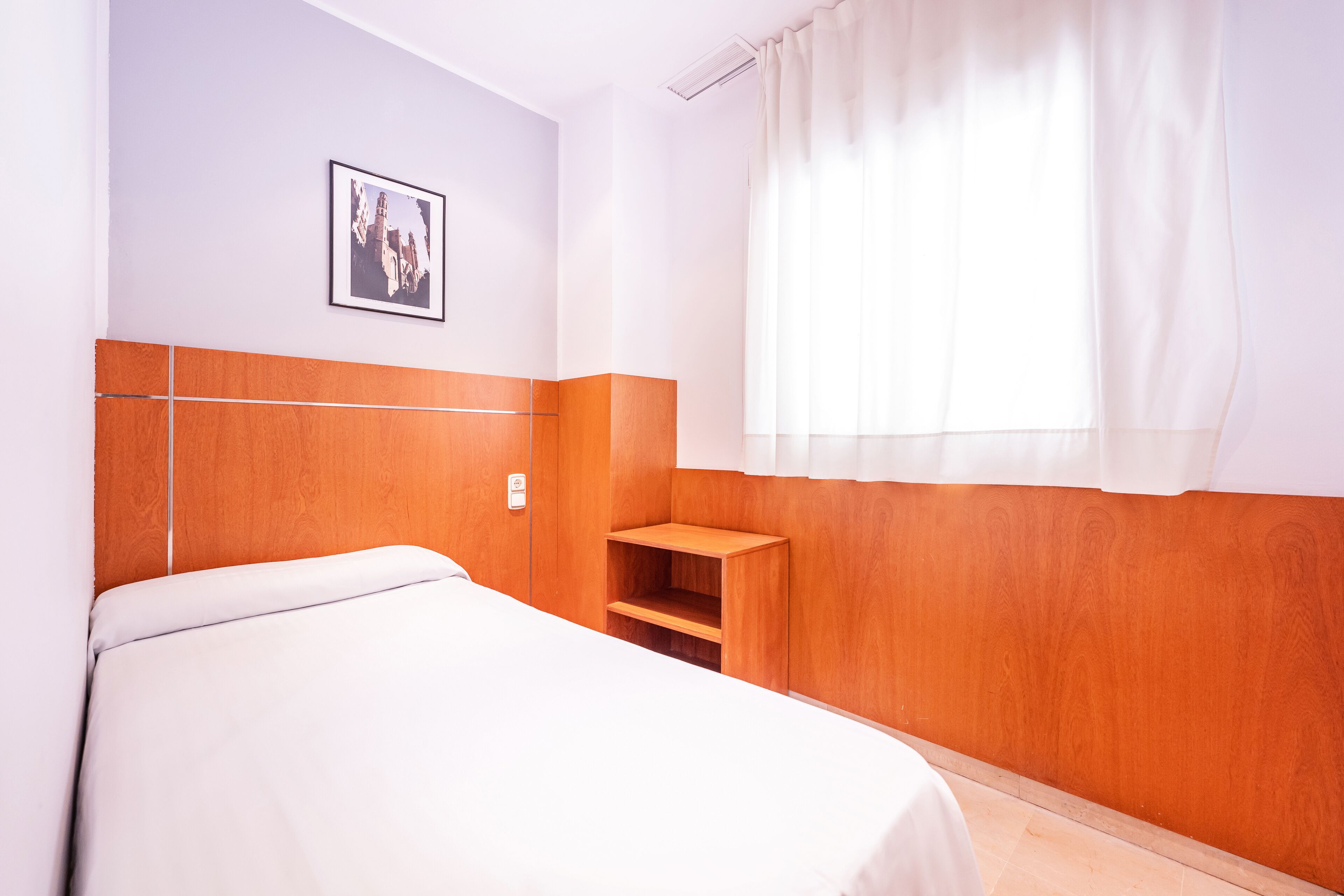 Ein Hotelzimmer mit einem Bett, einem Holz-Kopfteil, einer kleinen Holzhilfe, einem Fenster mit weißen Vorhängen und einem gerahmten Bild an der Wand.