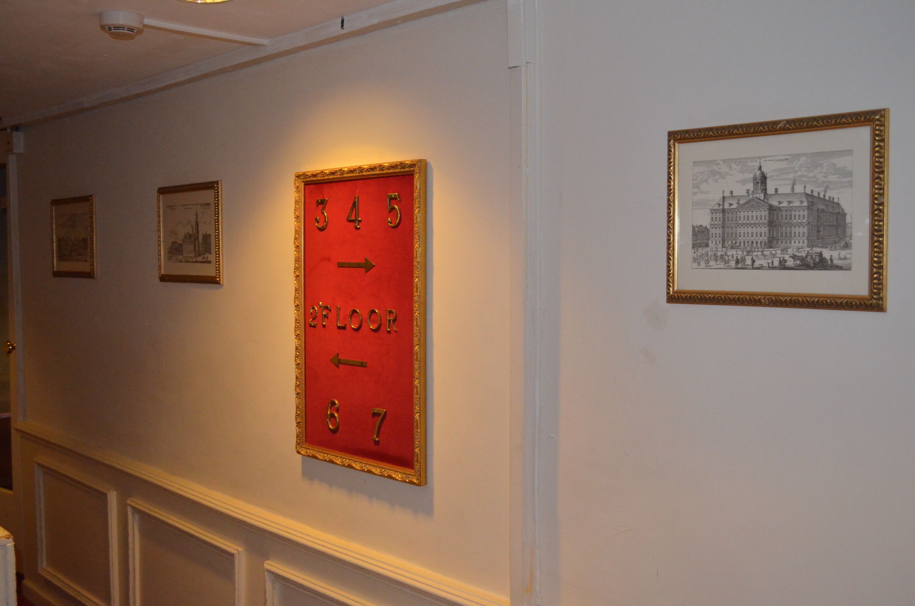 Ein Flur mit einem roten Hinweisschild, das die Zimmernummern 345 und 347 anzeigt, und einer schwarz-weißen Illustration eines historischen Gebäudes in einem Bilderrahmen an der Wand.