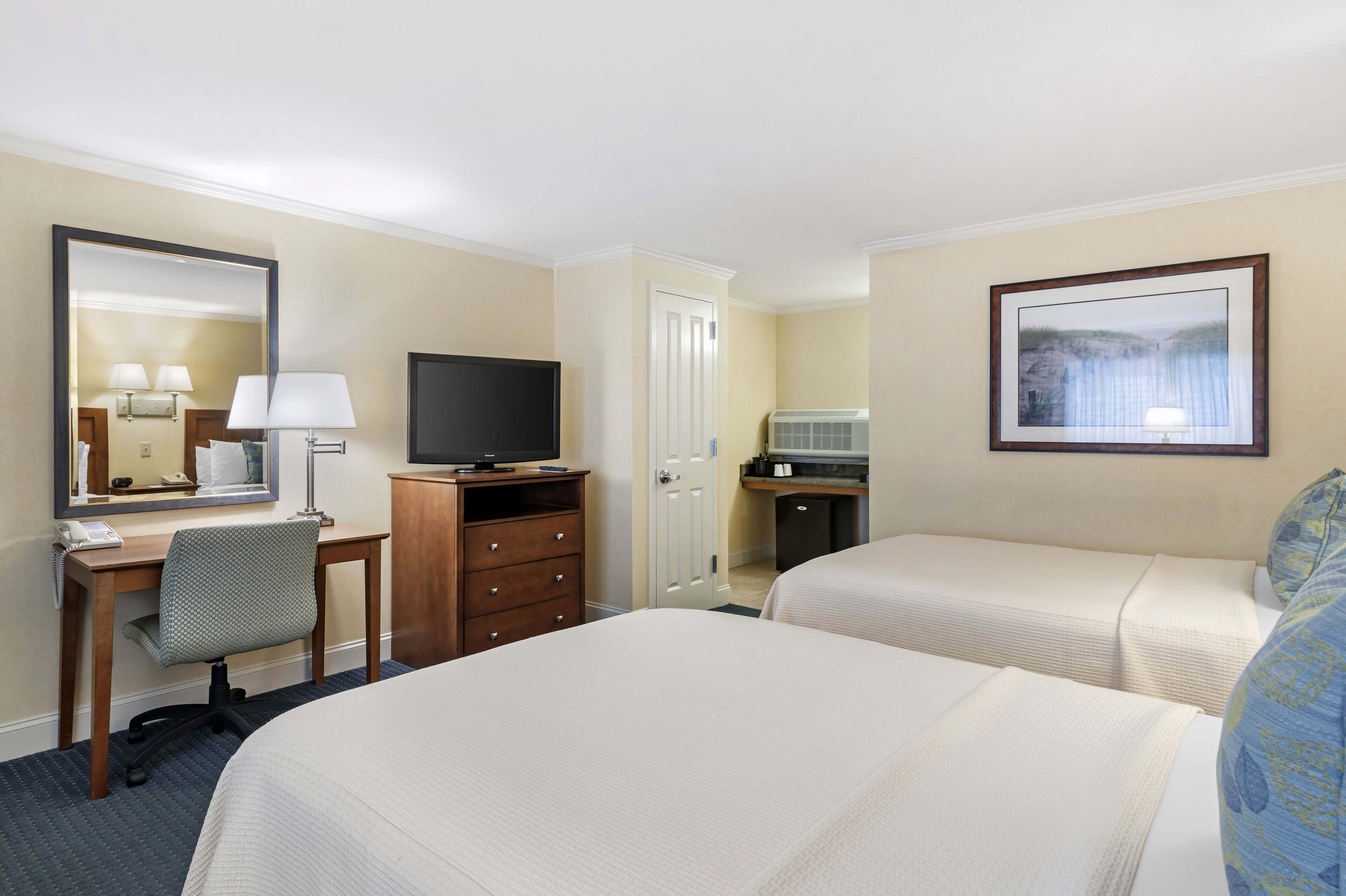 Habitación de hotel con dos camas, un escritorio con silla, televisor, lámpara de escritorio, espejo y un cuadro en la pared.