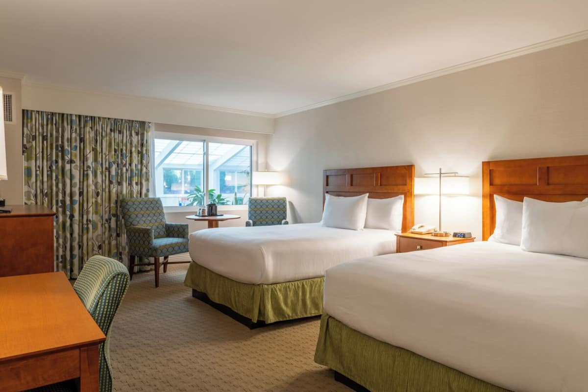 A hotel room with two beds, a desk, a chair, a lamp, and a window with curtains.