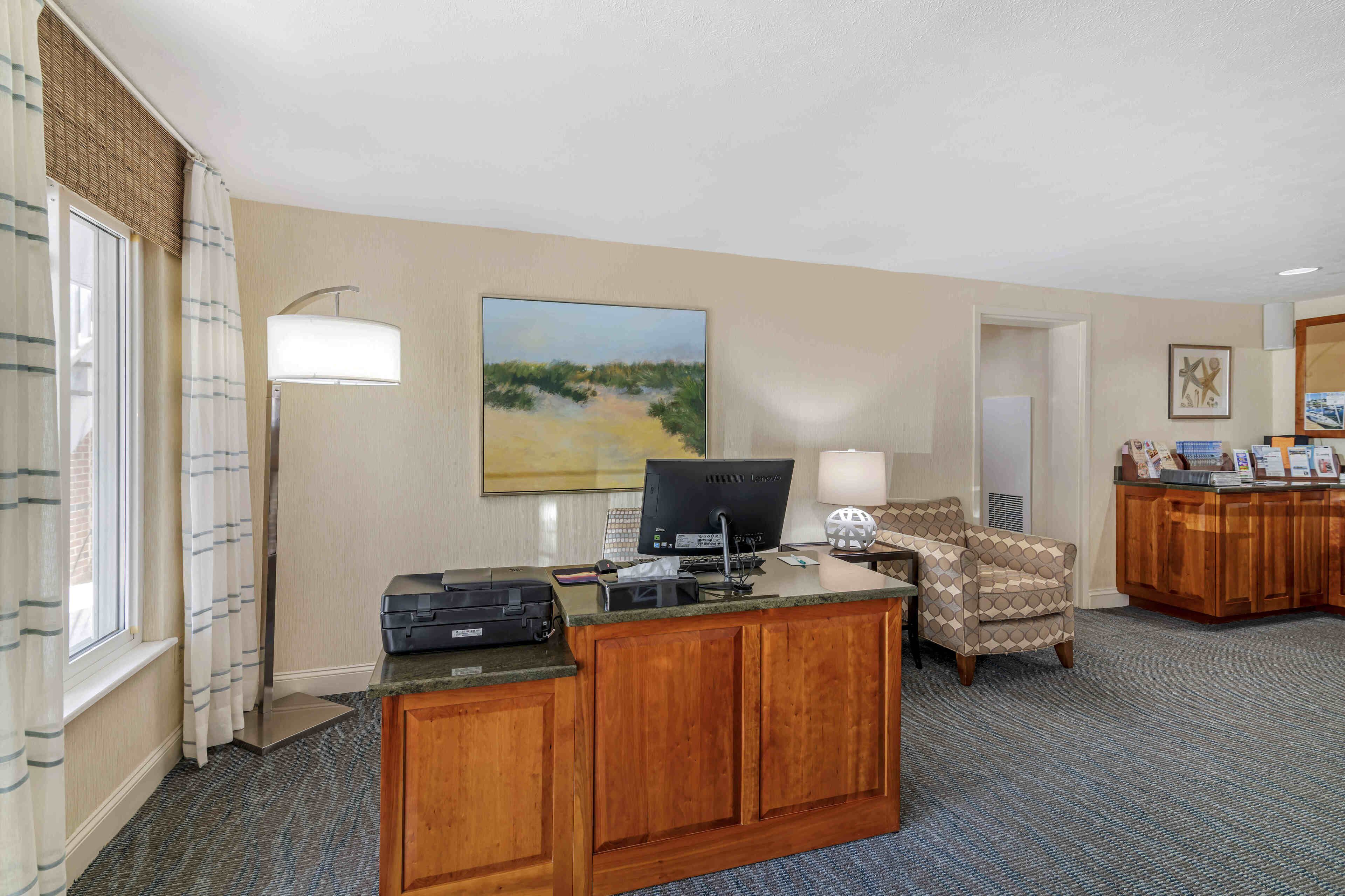 A hotel room with a wooden desk, a computer, a lamp, a chair, and a painting on the wall.