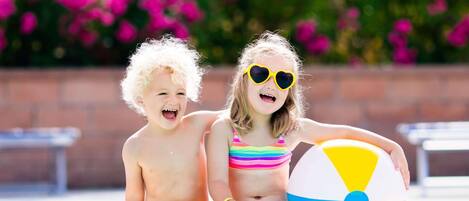 Two children sitting by a pool with a beach ball, surrounded by a brick wall and pink flowers.