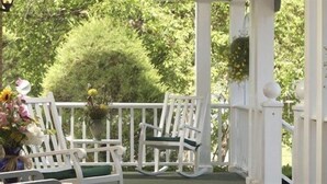 A white wooden porch with two rocking chairs and a small table, surrounded by lush greenery and flowering plants. - Juniper Hill Inn (Ogunquit)