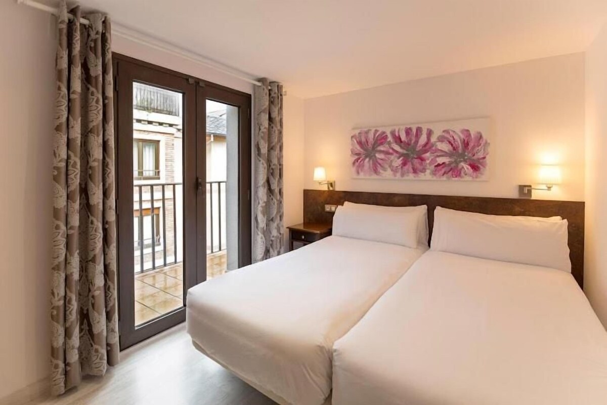 Habitación de hotel con cama doble, mesita de noche, un cuadro y vista a través de un ventanal.