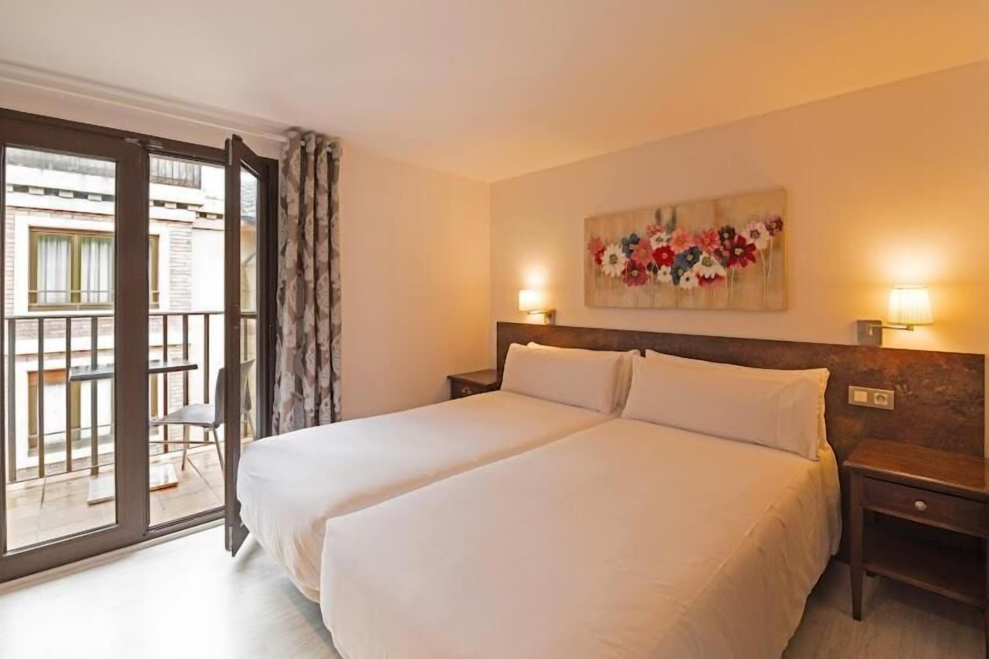 A double bed with white linens, a wooden headboard, a nightstand with a lamp, and a balcony with a chair.