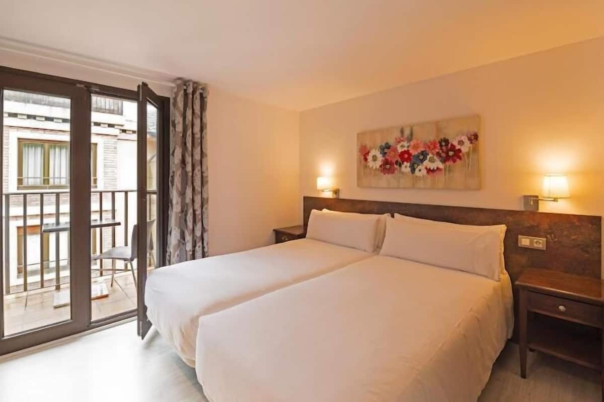 Un lit double avec du linge de lit blanc, une tête de lit en bois, une table de chevet avec une lampe et un balcon avec une chaise.