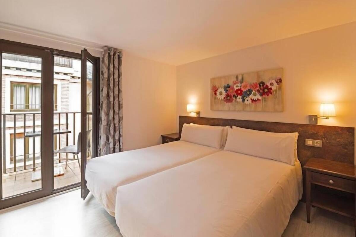 Cama doble con ropa blanca, cabecera de madera, mesita de noche con lámpara y balcón con una silla.