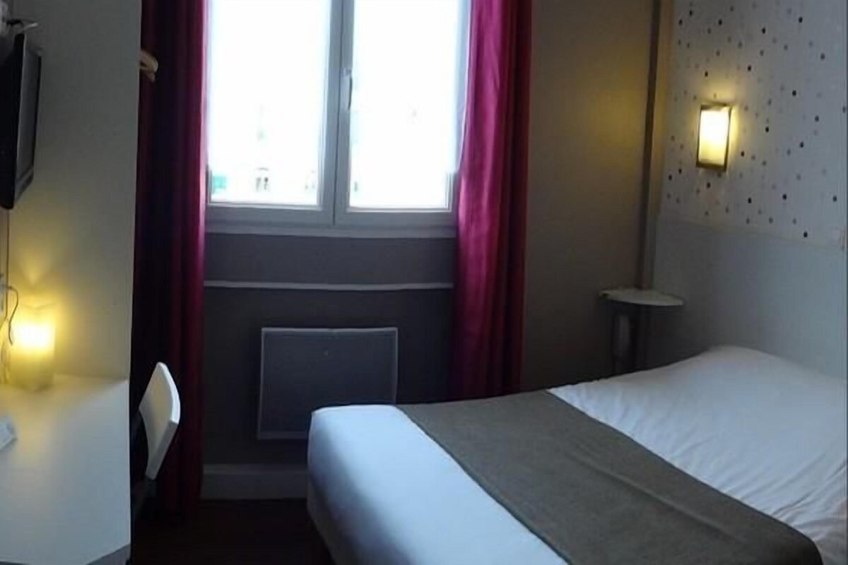 A hotel room with a bed, a desk, a chair, a TV, a window with curtains, and a wall-mounted light.
