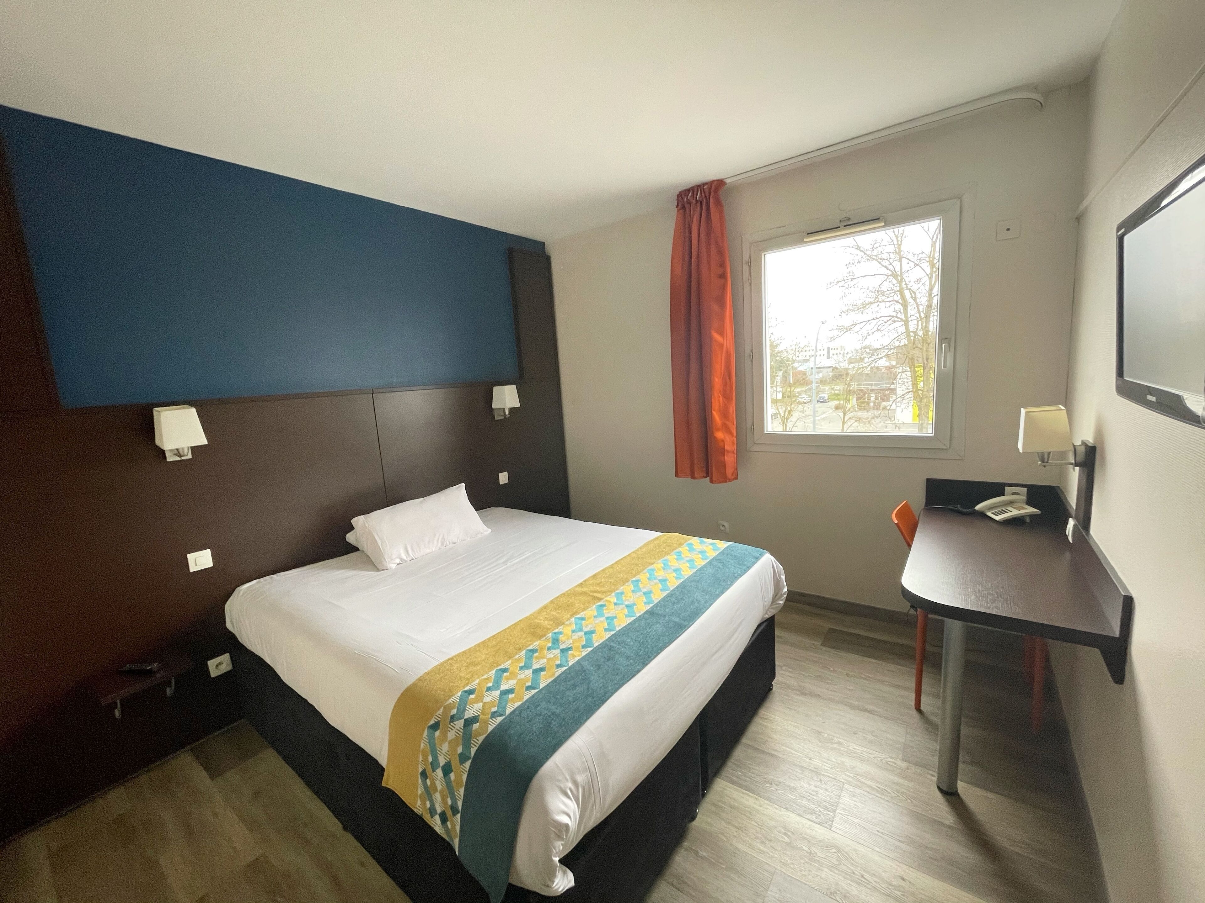 Ein Hotelzimmer mit einem Bett, einem Schreibtisch, einem Stuhl, einem fenster mit Vorhängen und einem an der Wand befestigten Fernseher.