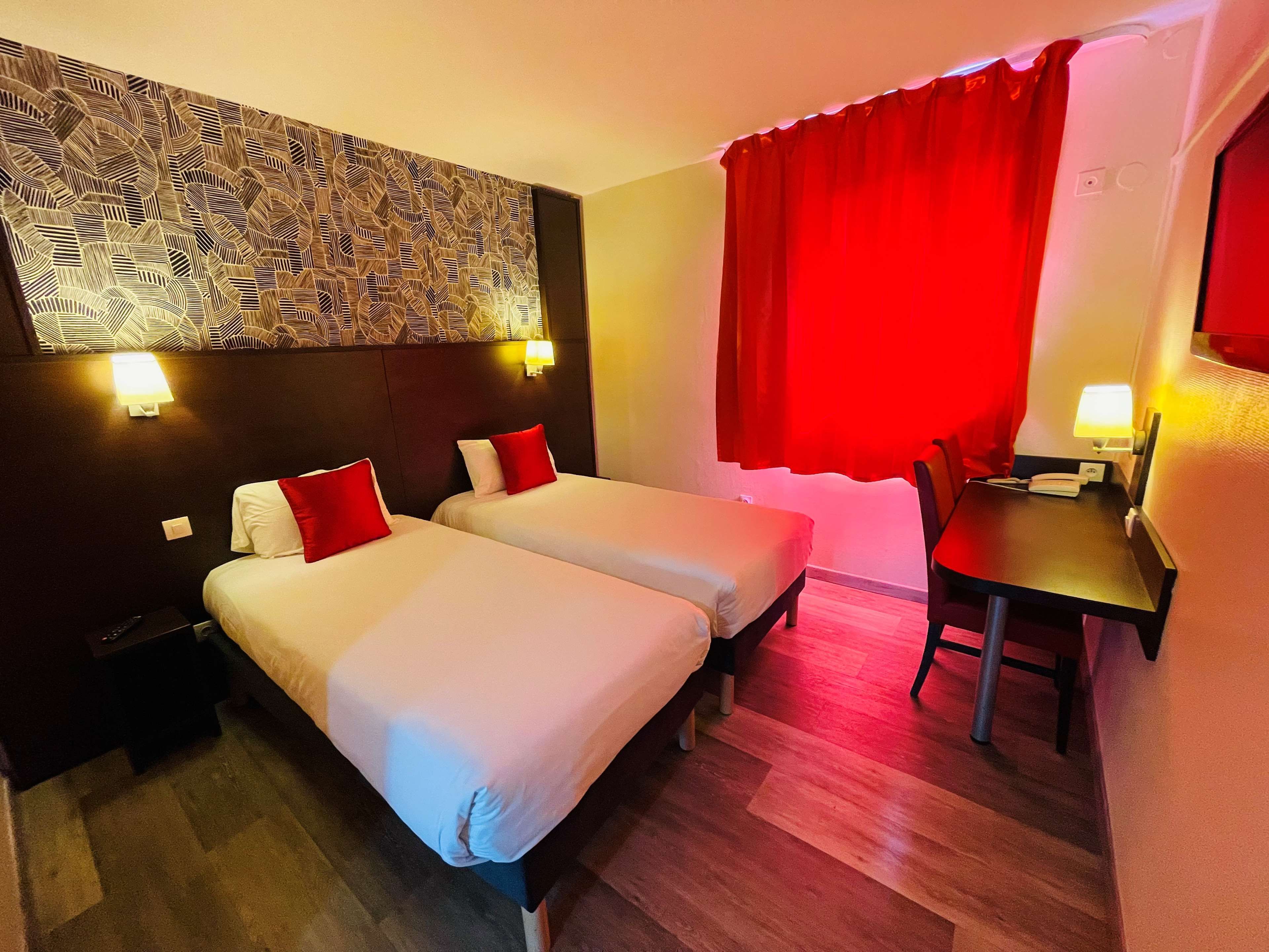 Une chambre d’hôtel avec deux lits, un bureau, une chaise, un rideau rouge et un papier peint à motifs.