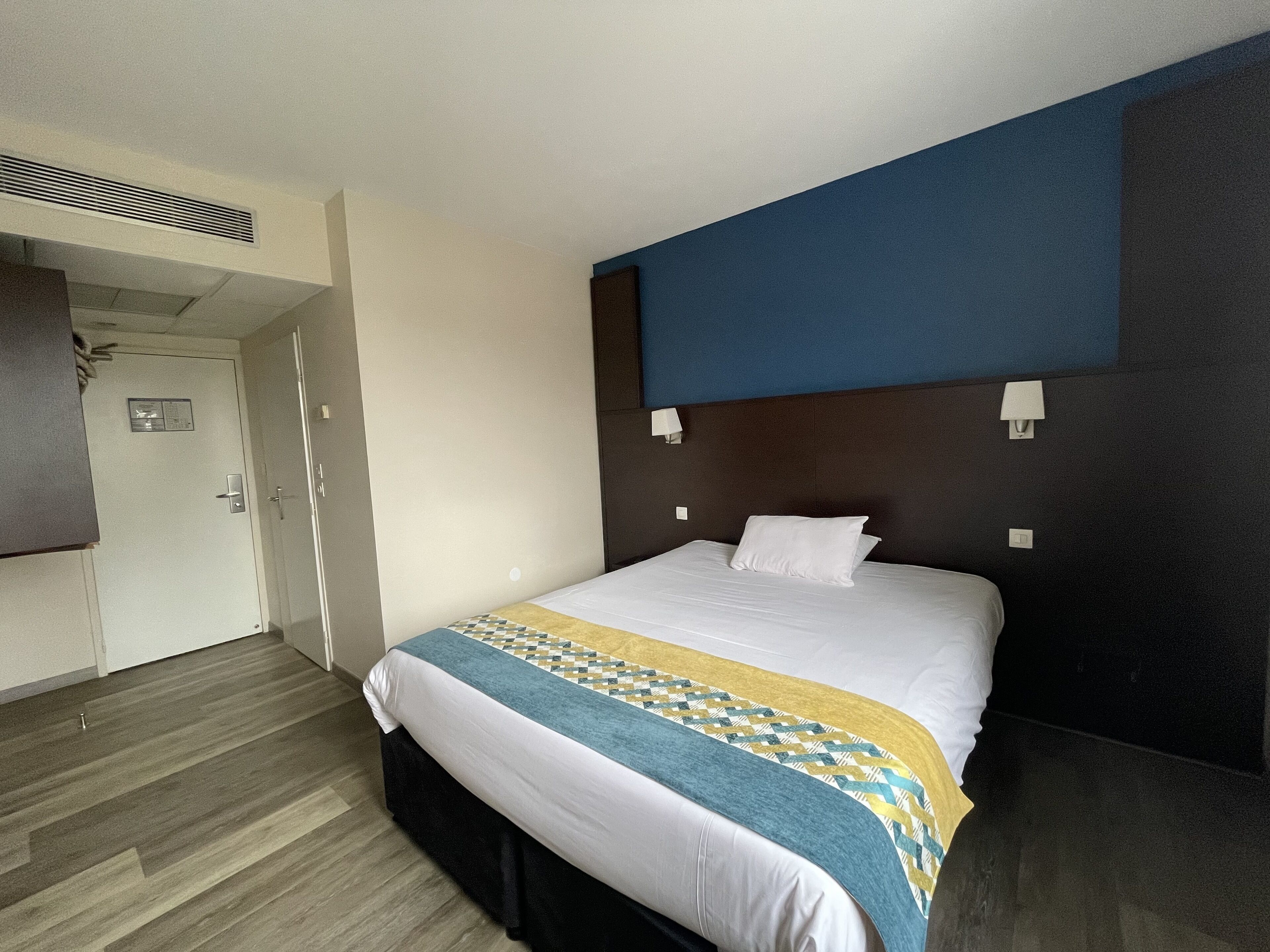 A hotel room with a bed, a door, a wall-mounted light, and a vent.
