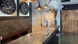 Een moderne receptiezone met twee grote wandklokken, een houten bank, een balie met handdesinfectiemiddel en een glas, en hanglampen.