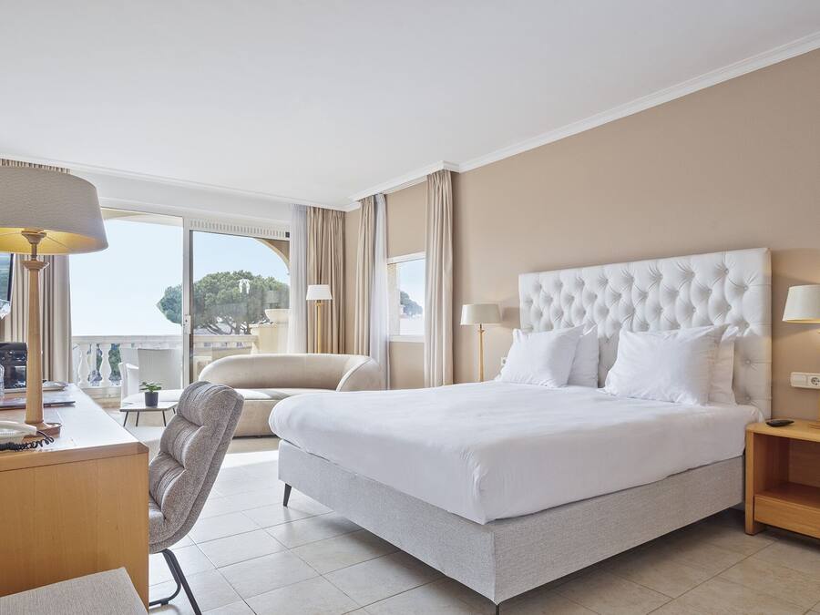 Ein Hotelzimmer mit einem großen Bett, einem Schreibtisch mit Lampe, einem Sessel und Blick ins Freie.