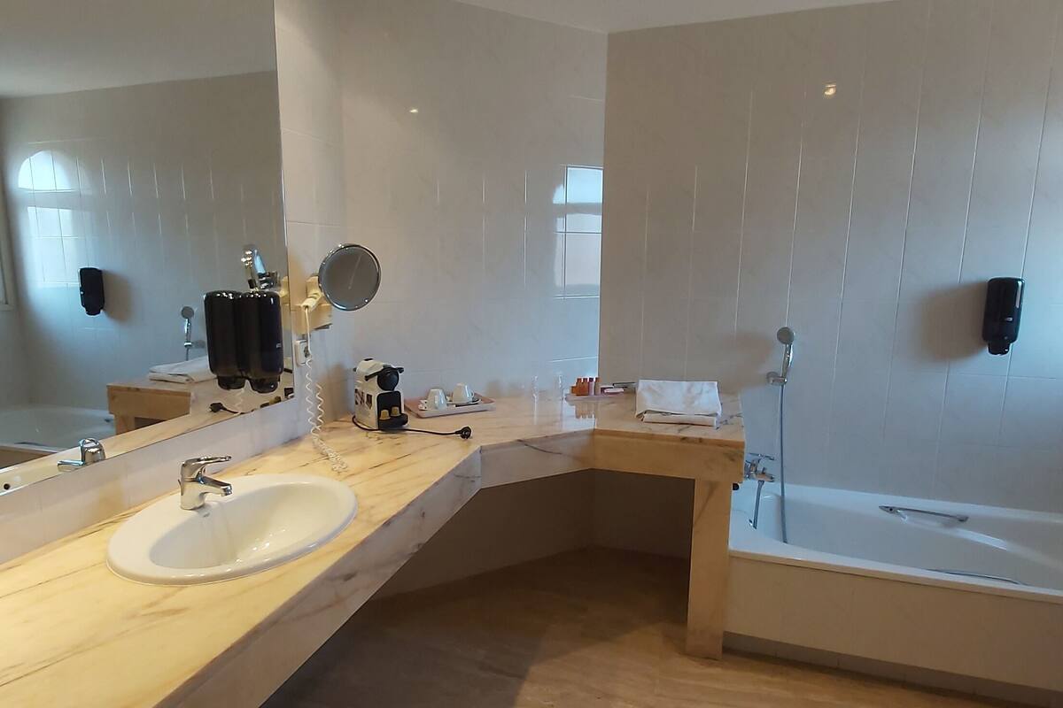 A modern bathroom with a large mirror, two sinks, a bathtub, and a window.
