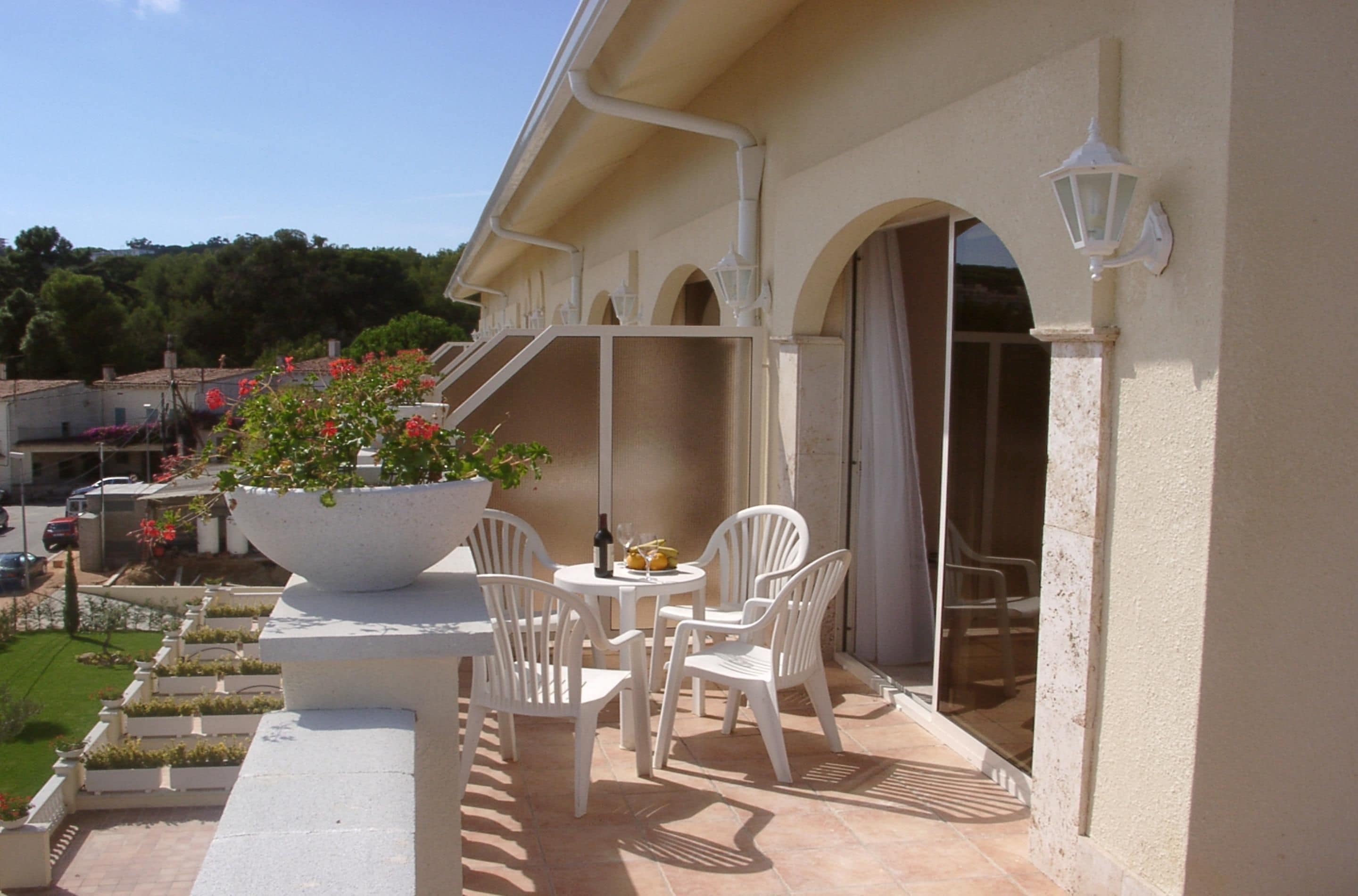 Un patio al aire libre con sillas y una mesa blancas, una maceta grande con flores y vistas del entorno.