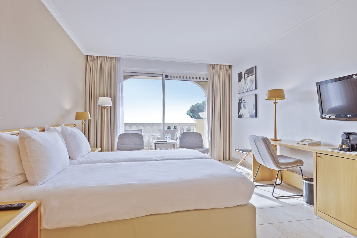 Ein Hotelzimmer mit einem großen Bett, einem Schreibtisch mit Stuhl, einem Fernseher und einem Balkon mit Tisch und Stühlen.