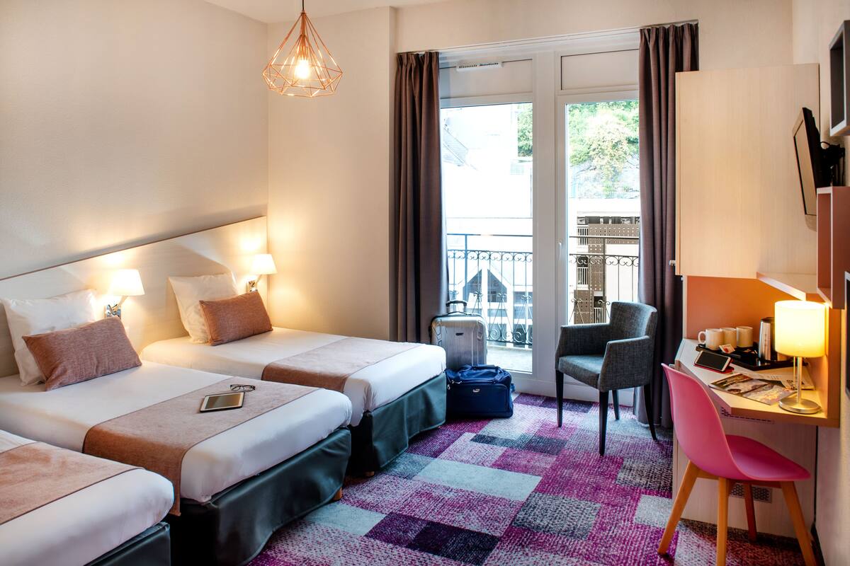 A hotel room with two beds, a desk with a chair, a suitcase, and a balcony with a railing.