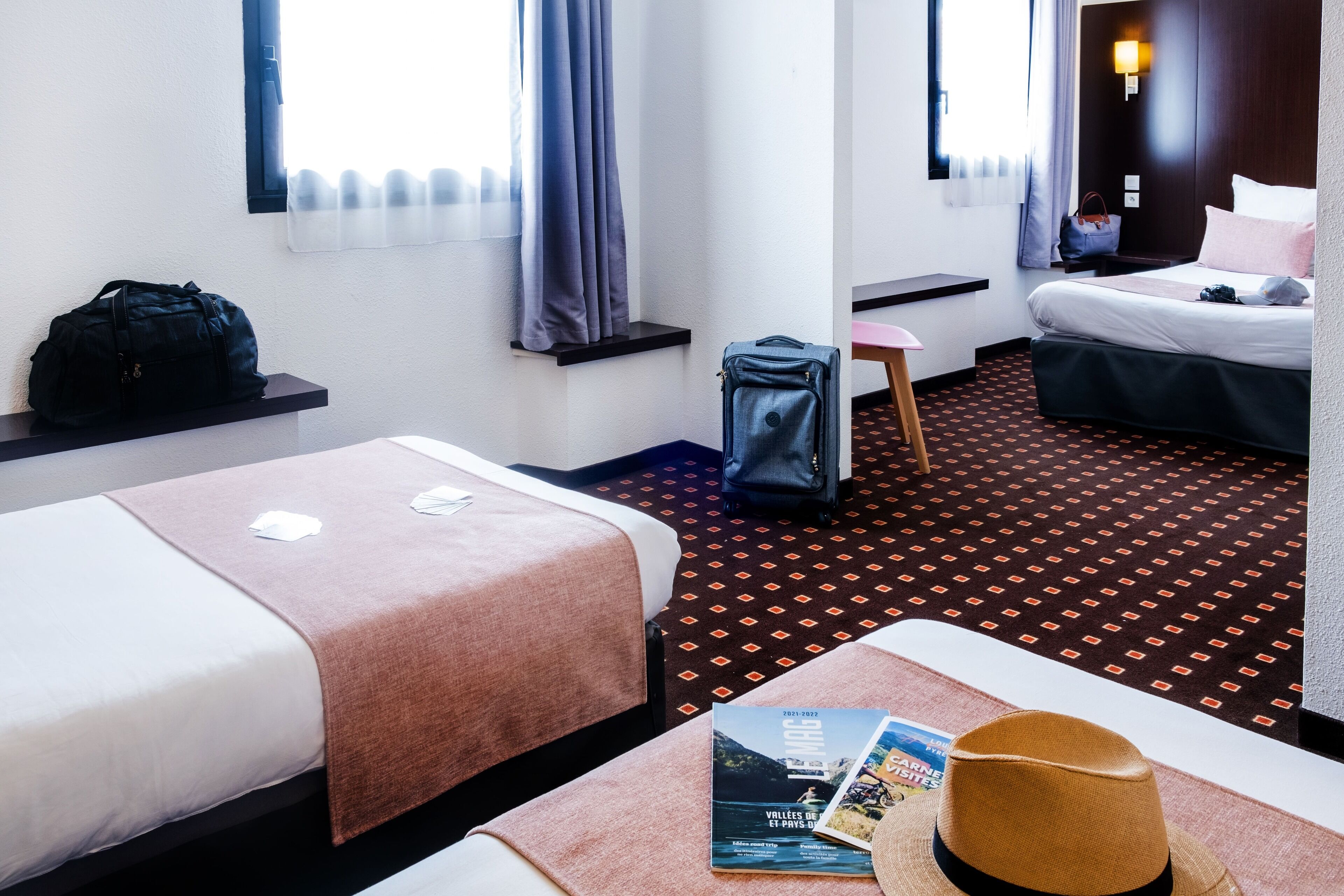 A hotel room with two beds, a desk, a chair, a suitcase, a handbag, and a magazine on the bed.