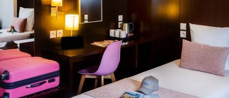 A hotel room with a bed, a desk, a chair, a pink suitcase, and a cap on the bed.