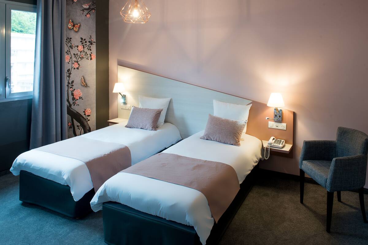 A hotel room with two beds, a chair, a nightstand with a lamp, and a wall-mounted phone.