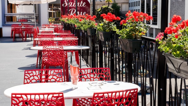 Outdoor seating with red chairs and white tables, a sign for 'Sainte Rose', and a building with 'HOTEL ROC DE MASSABIELLE' in the background. - Hotel Sainte Rose (Lourdes)