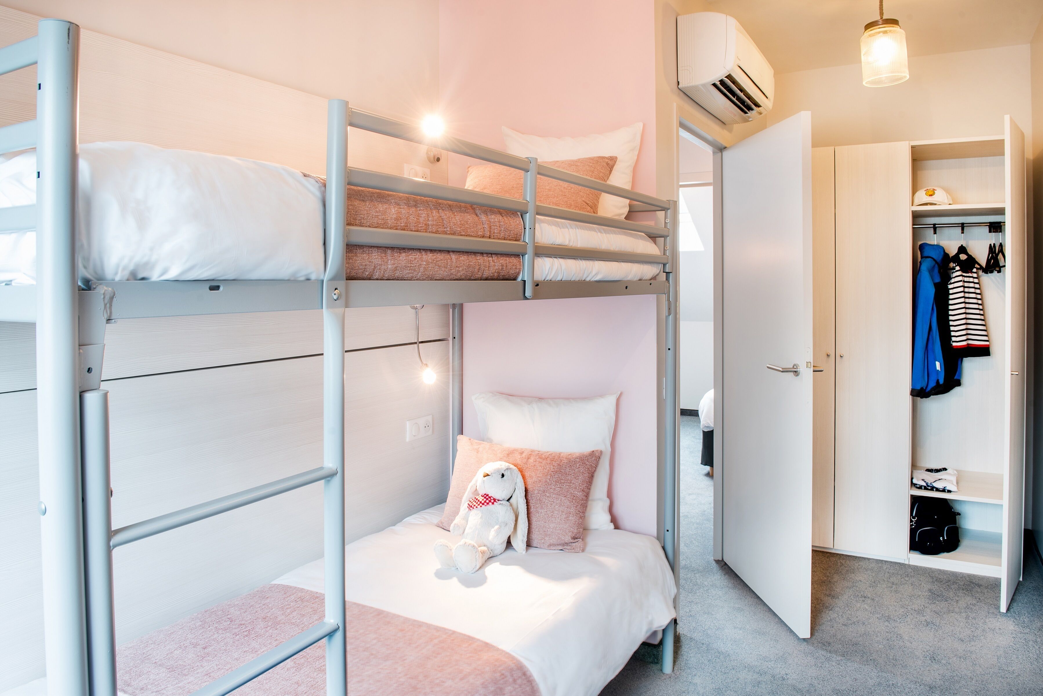 Een stapelbed met een knuffel, een kledingkast met daarin hangende kleding, en een zichtbare airconditioning.