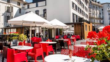 Un café en plein air avec des tables et des chaises rouges et blanches, des parasols blancs et des plantes en pot.