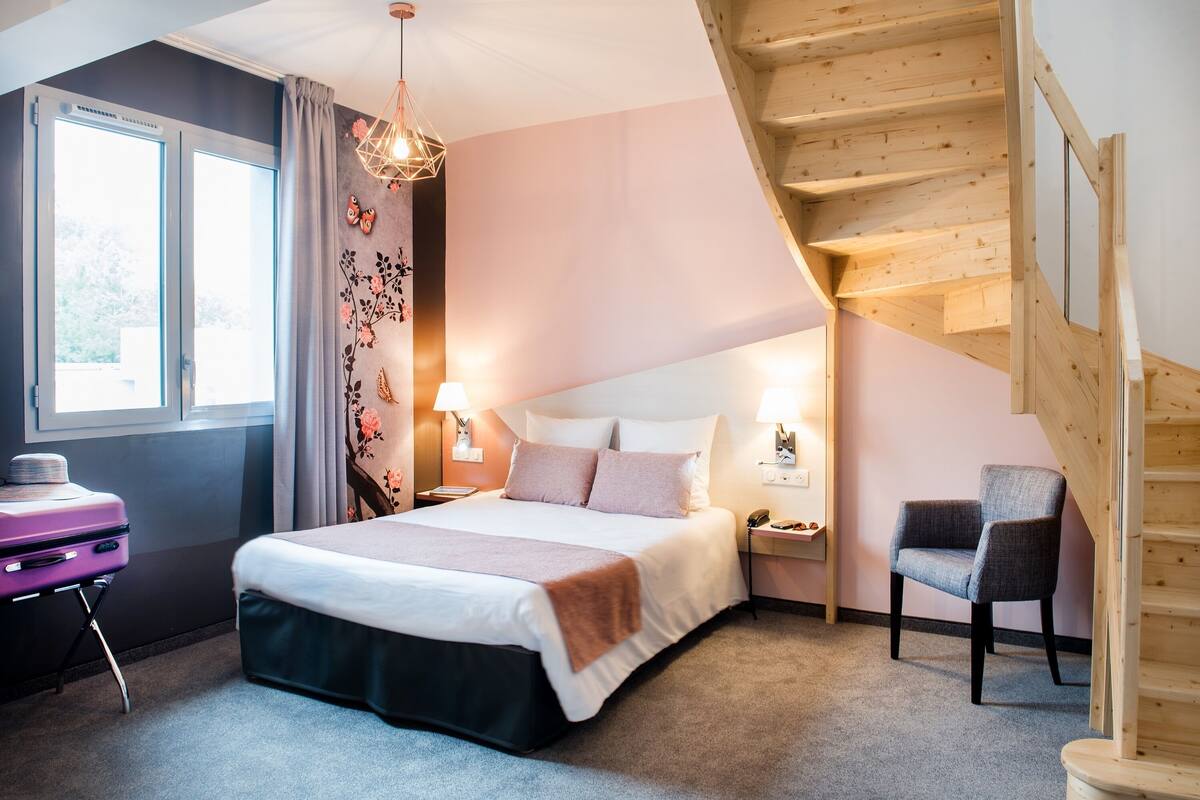 A bedroom with a bed, bedside lamps, a chair, a suitcase, and a wooden staircase.