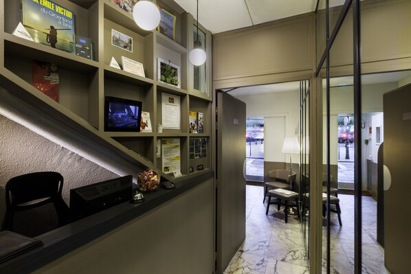 A reception area with a counter, seating, and a glass door leading to a lobby. - Contact Hotel Alizé Montmartre (Paris)