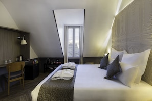 A modern hotel room with a large bed, a desk, and a chair. There is a window with curtains, a lamp, and a decorative headboard. - Contact Hotel Alizé Montmartre (Paris)