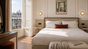 A hotel room with a large bed, a bedside table with a lamp, a window with a view of buildings, and a painting on the wall. - Hôtel Beauvoisin (Paris)