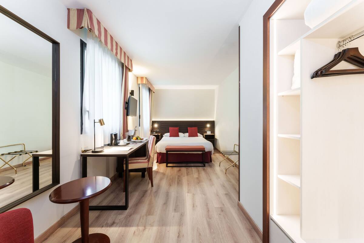 Una habitación de hotel con cama, mesitas de noche, un escritorio con silla, un espejo y un armario.