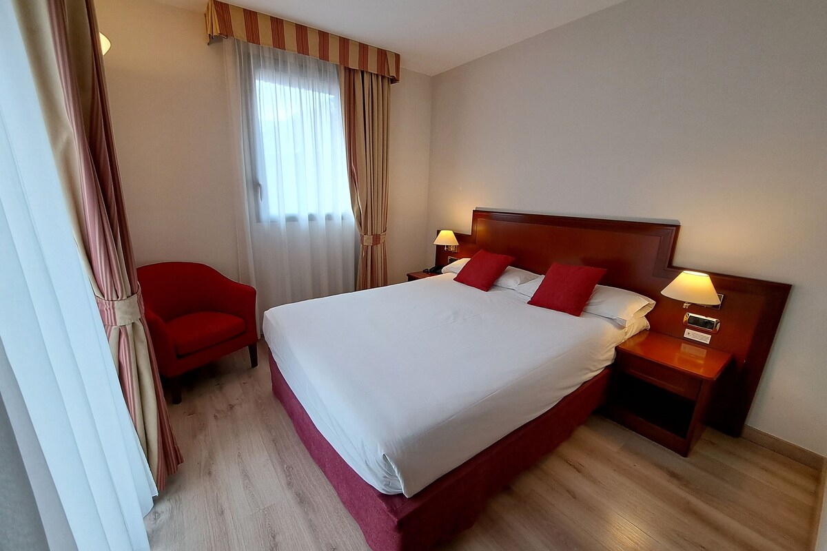 Ein Hotelzimmer mit einem großen Bett, zwei Nachttischen inklusive Lampen, einem roten Sessel und einem Fenster mit Vorhängen.