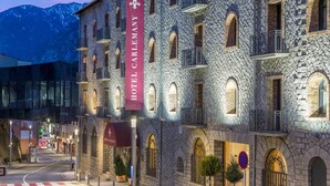 Un edificio histórico de piedra con varios balcones y ventanas arqueadas, un llamativo letrero rojo que dice "HOTEL CARLEMANY", y un paisaje montañoso de fondo.