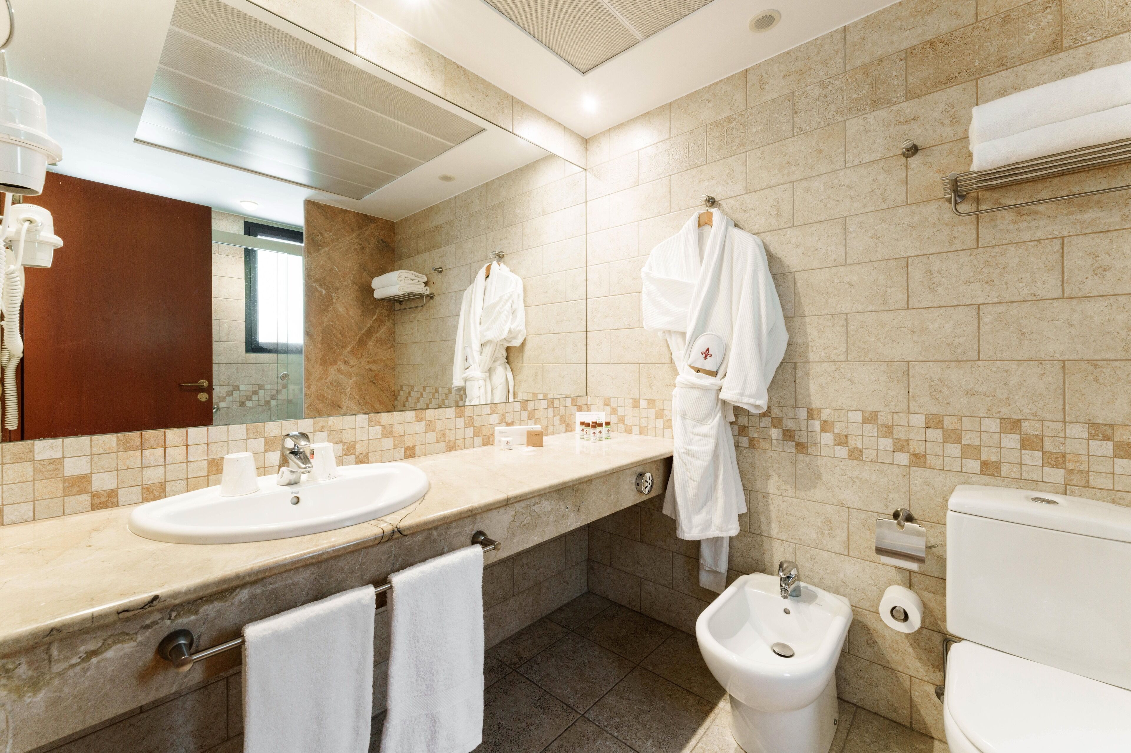 Une salle de bain moderne avec un grand miroir, deux lavabos, un bidet et un porte-serviettes avec des peignoirs.