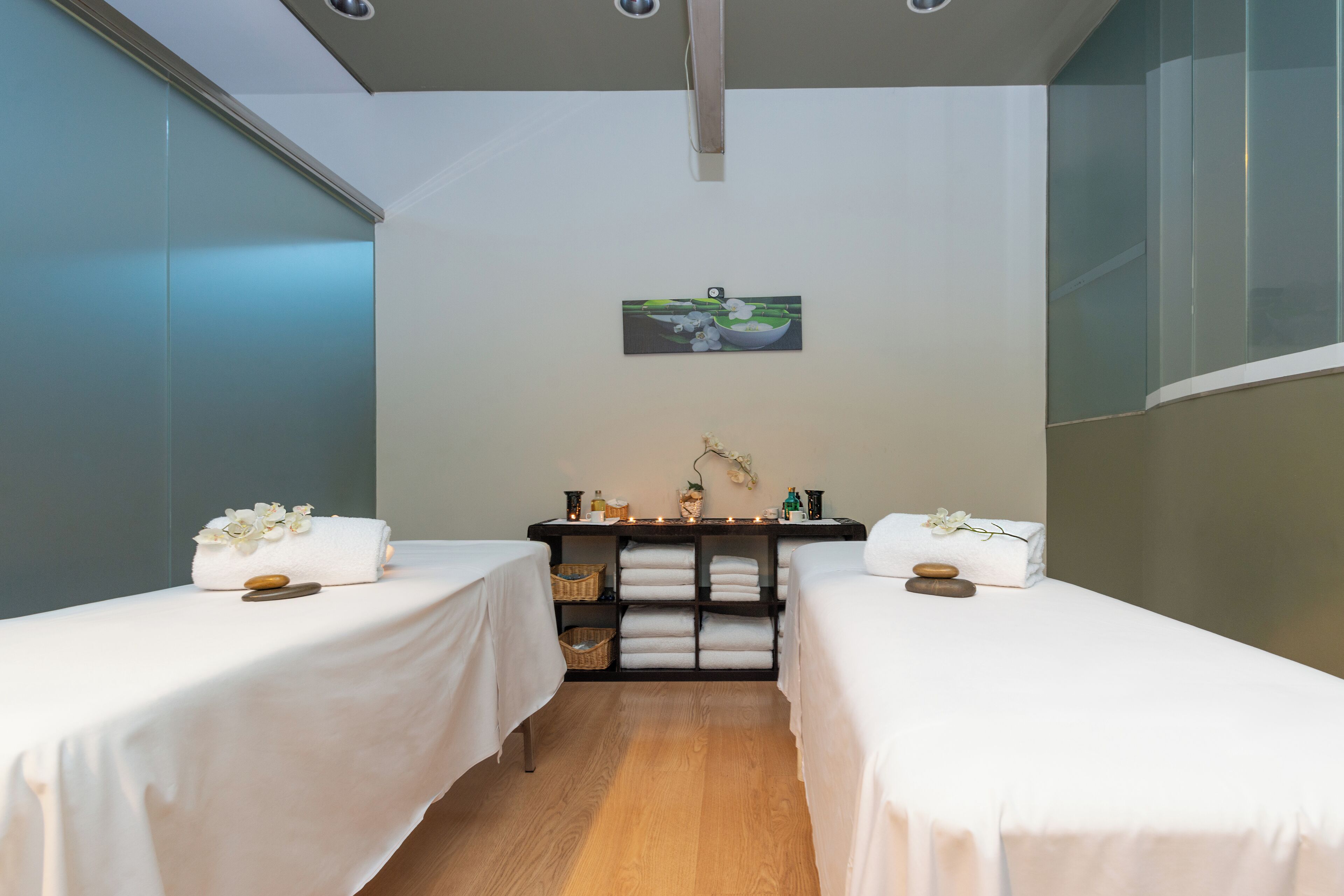 A spa room with two beds, a shelf with towels and toiletries, and a painting on the wall.