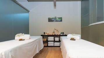 A spa room with two beds, a shelf with towels and toiletries, and a painting on the wall.