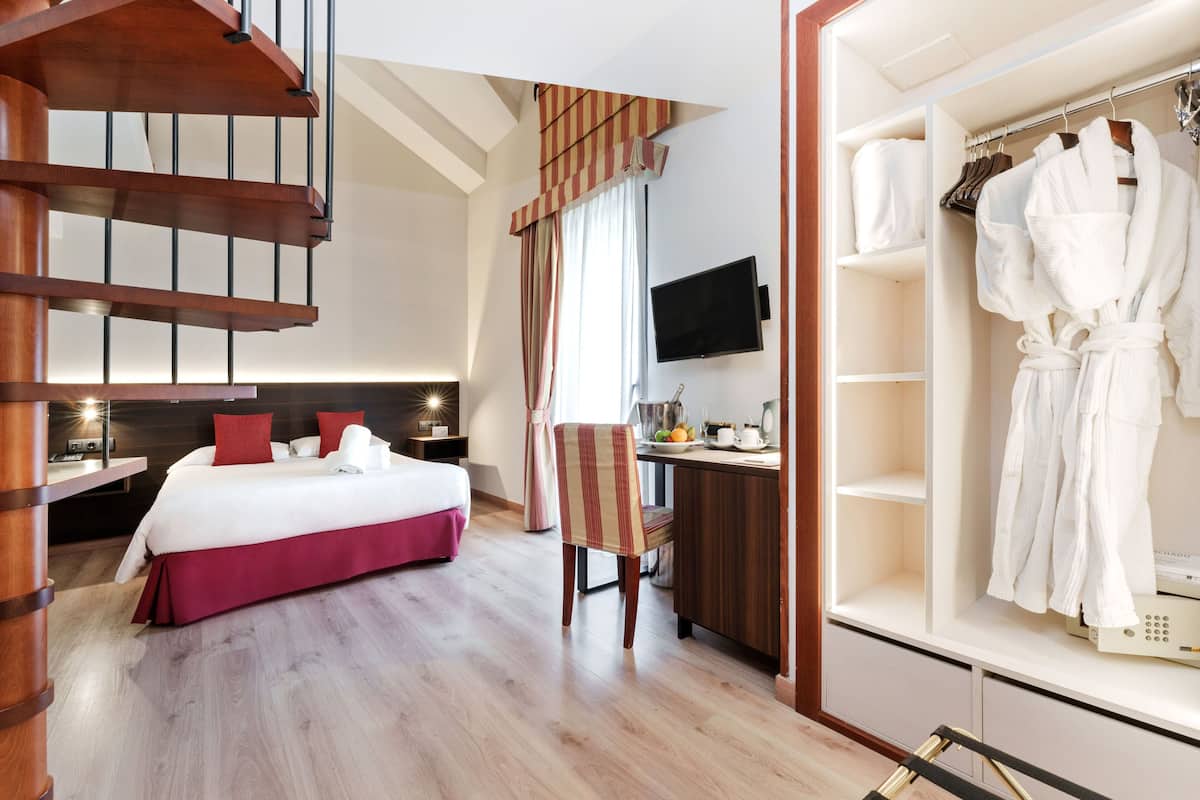 Habitación de hotel con una escalera de madera, una cama con ropa de cama roja, un escritorio con silla, un televisor y un vestidor con batas.