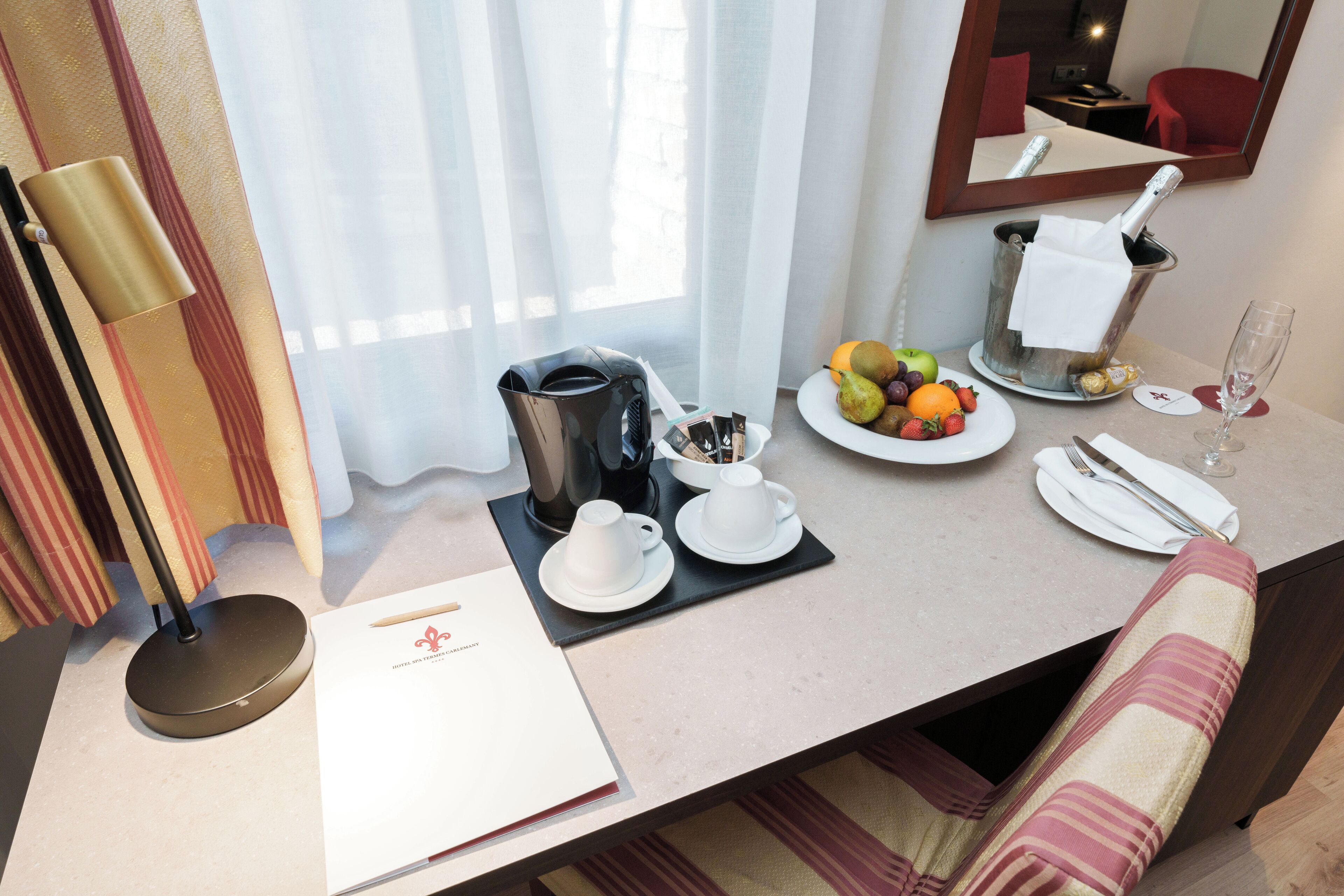 Habitación de hotel con una mesa de desayuno ordenada, que incluye una cafetera, tazas de té, fruta y una copa de vino.