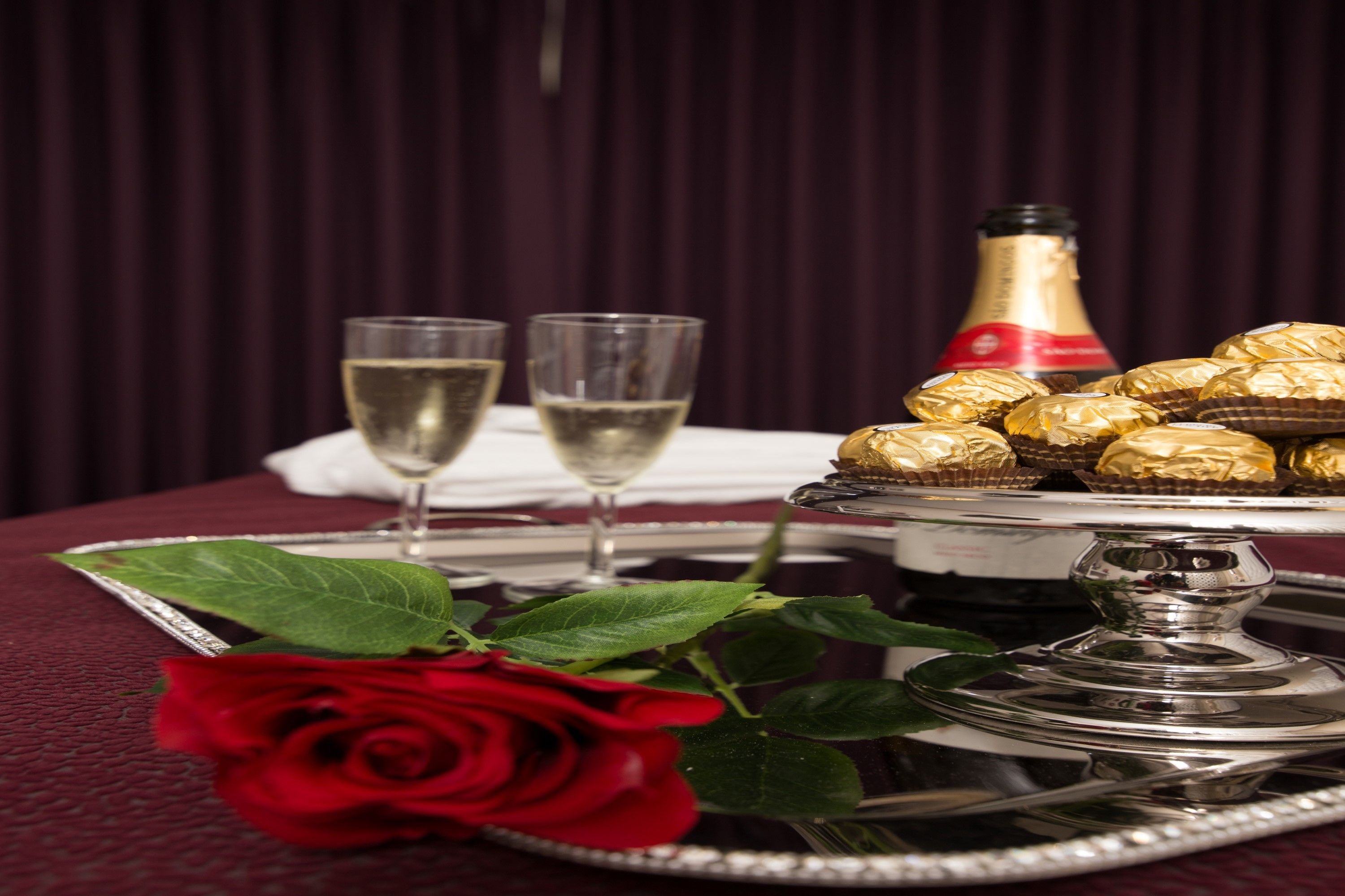 A table with a silver tray holding chocolates, two glasses of champagne, and a red rose.
