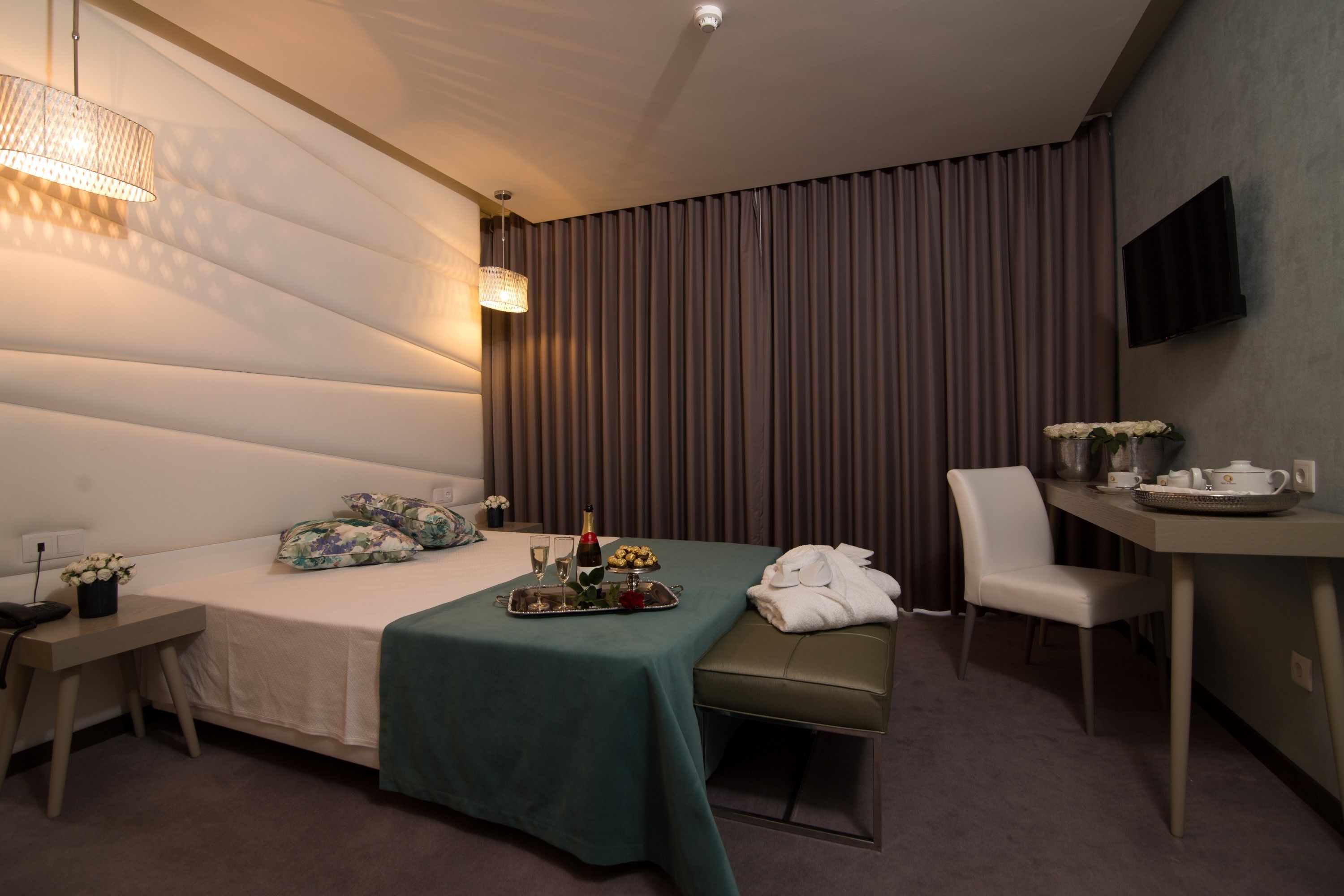 Habitación de hotel con cama, mesita de noche, un pequeño escritorio, una silla, televisor montado en la pared y ventana con cortinas.