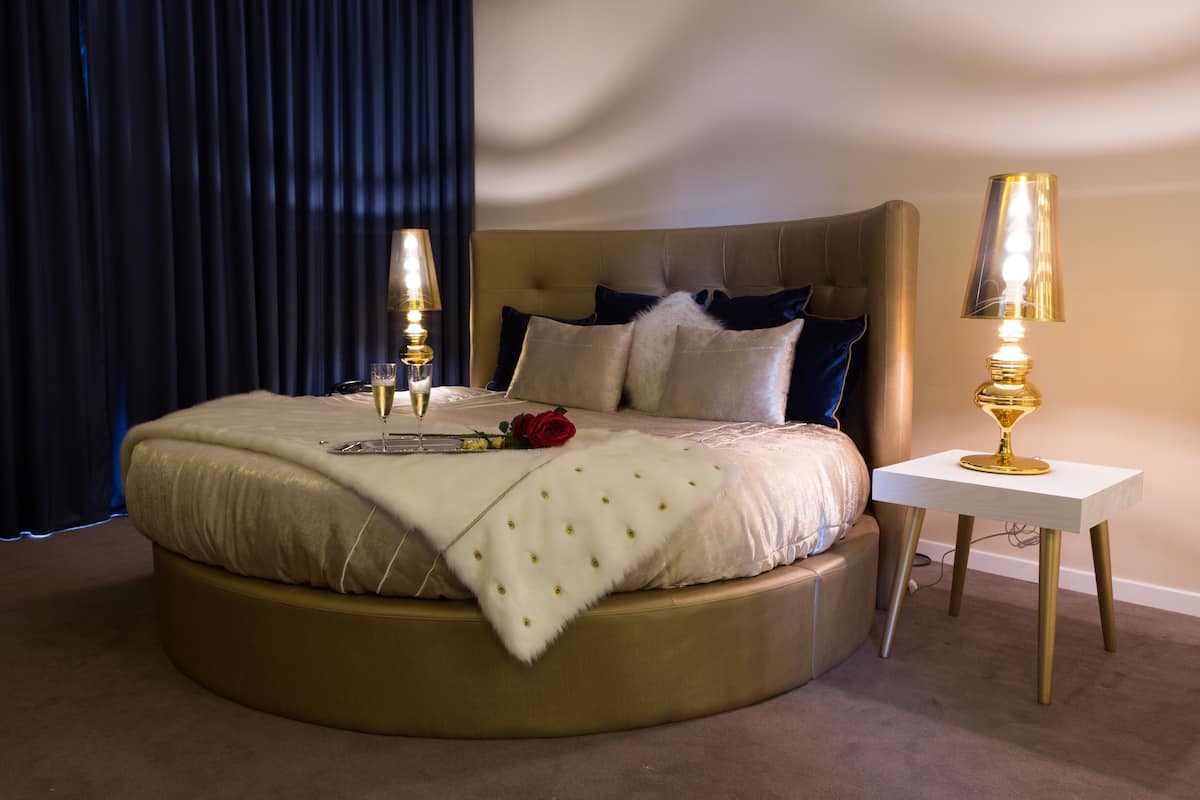 Een rond bed met een beige hoofdbord, twee bedlampen, een roos en een glas wijn op het nachtkastje.