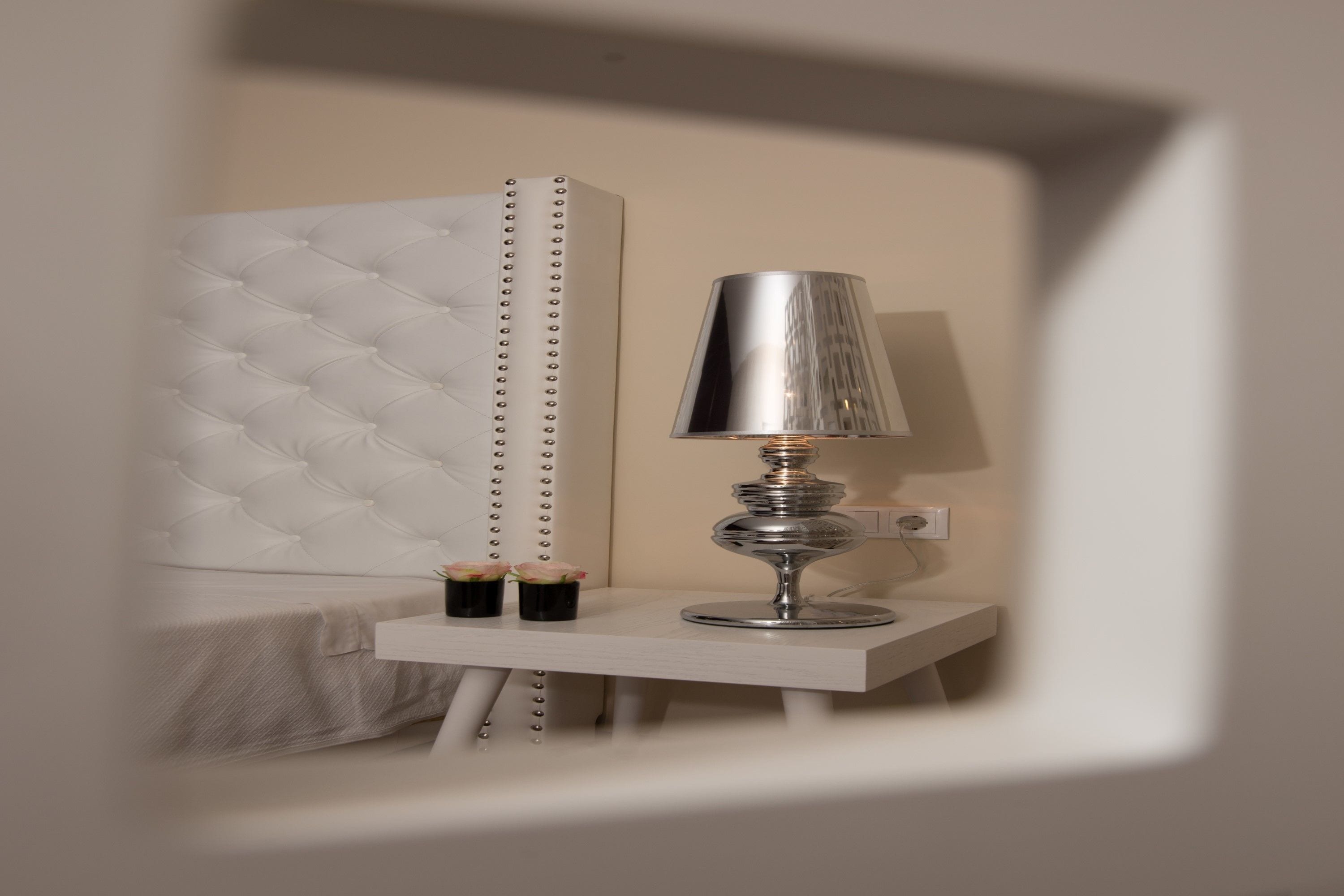 A bedside table with a lamp and two small cups, a tufted headboard, and a white wall.