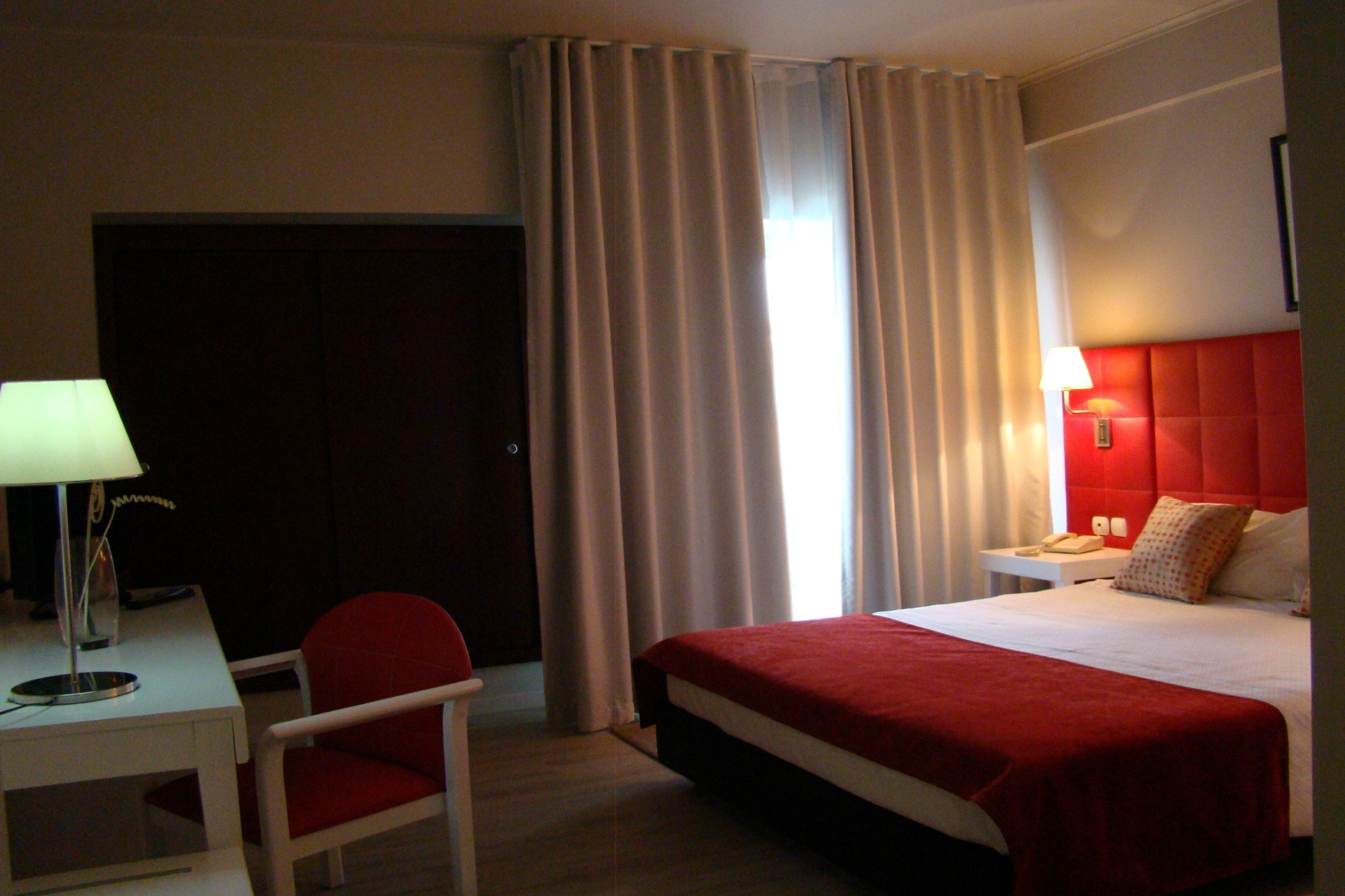 A hotel room with a bed, a desk with a lamp, a chair, a nightstand with a lamp, and a red headboard.
