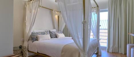 A four-poster bed with white bedding and blue pillows, a glass coffee table, and a view of the outdoors.